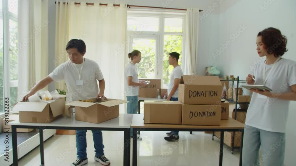 Medium long shot of young female volunteer keeping count of donation boxes using digital tablet in fund with other members of team Medium long shot of young female volunteer keeping count of donation boxes using digital tablet in fund with other members of team