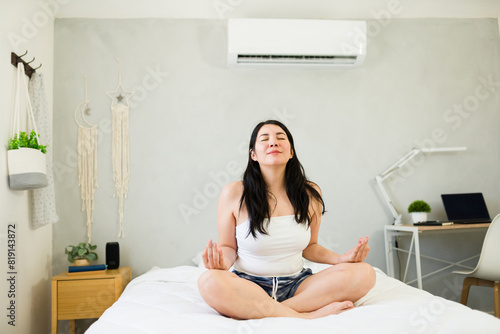 Content woman meditates on a bed under a modern mini-split ac unit, enjoying a cool and comfortable indoor breeze during summer