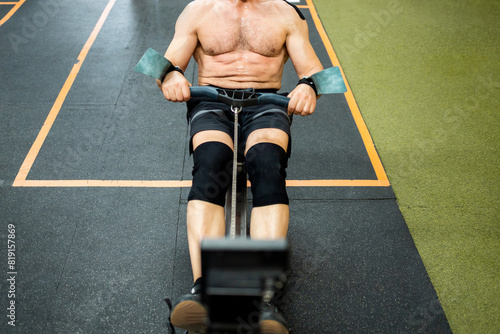 An unrecognizable senior man performs shirtless physical exercise inside a gym. The male is working his back with an air rowing machine. Rowing air machine concept.