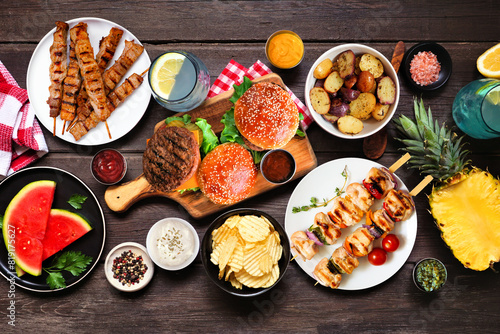 Summer BBQ food table scene. Hamburgers, meat skewers, potatoes, fruit and snacks. Top view on a dark wood background.