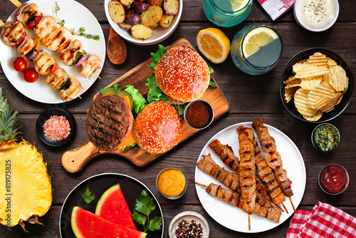 Summer BBQ food table scene. Hamburgers, meat skewers, potatoes, fruit and snacks. Overhead view on a dark wood background.