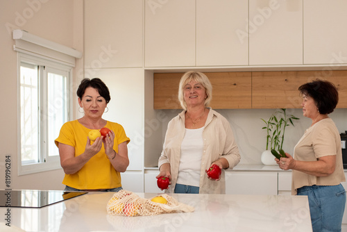 Three energetic mature women engage in conversation while holding fresh vegetables in a bright, modern kitchen. They seem relaxed and happy, sharing moments of friendship and domestic life.