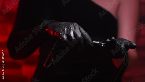 Close-up of female hands in black gloves holding a BDSM whip on a red background. BDSM is an outfit for adult sex games. Sex shop Adult store
