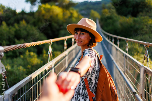 Girl with a backpack. outdoor portrait of a young woman walking on a suspension bridge over a river, the girl turns and extends her hand to the camera as if she is following me.
