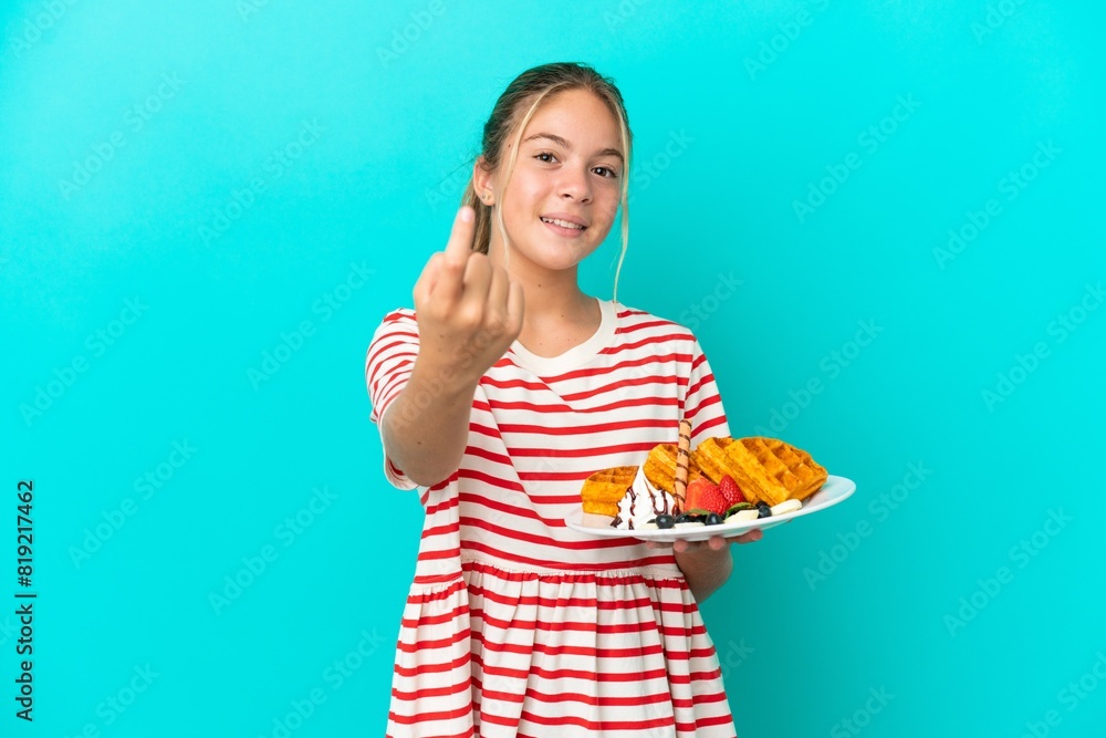 Little caucasian girl holding waffles isolated on blue background doing coming gesture Little caucasian girl holding waffles isolated on blue background doing coming gesture