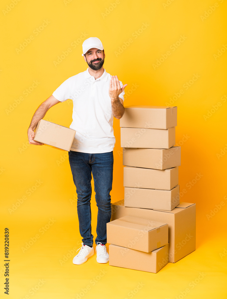 Full-length shot of delivery man among boxes over isolated yellow background inviting to come with hand. Happy that you came Full-length shot of delivery man among boxes over isolated yellow background inviting to come with hand. Happy that you came
