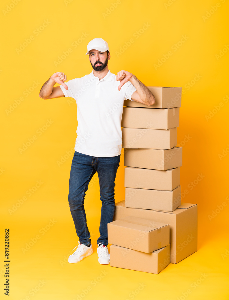 Full-length shot of delivery man among boxes over isolated yellow background showing thumb down Full-length shot of delivery man among boxes over isolated yellow background showing thumb down