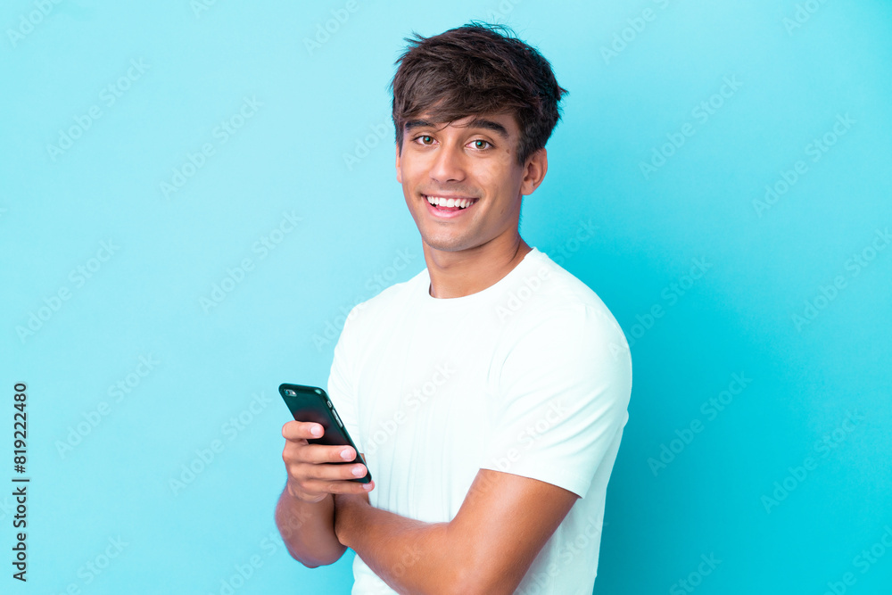 Young caucasian man isolated on blue background holding a mobile phone and with arms crossed Young caucasian man isolated on blue background holding a mobile phone and with arms crossed