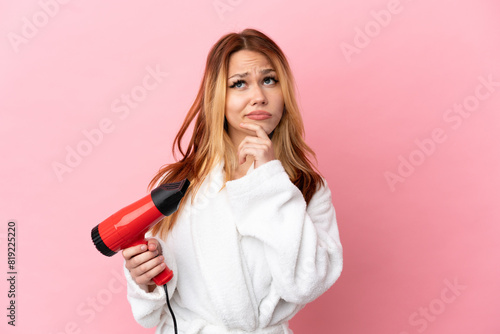 Teenager blonde girl holding a hairdryer over isolated pink background having doubts