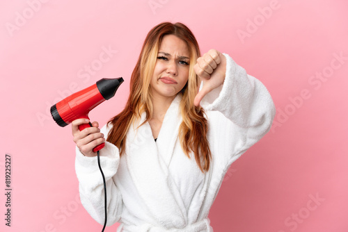 Teenager blonde girl holding a hairdryer over isolated pink background showing thumb down with negative expression