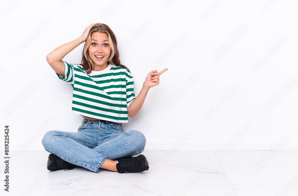 Young Russian girl sitting on the floor isolated on white background surprised and pointing finger to the side