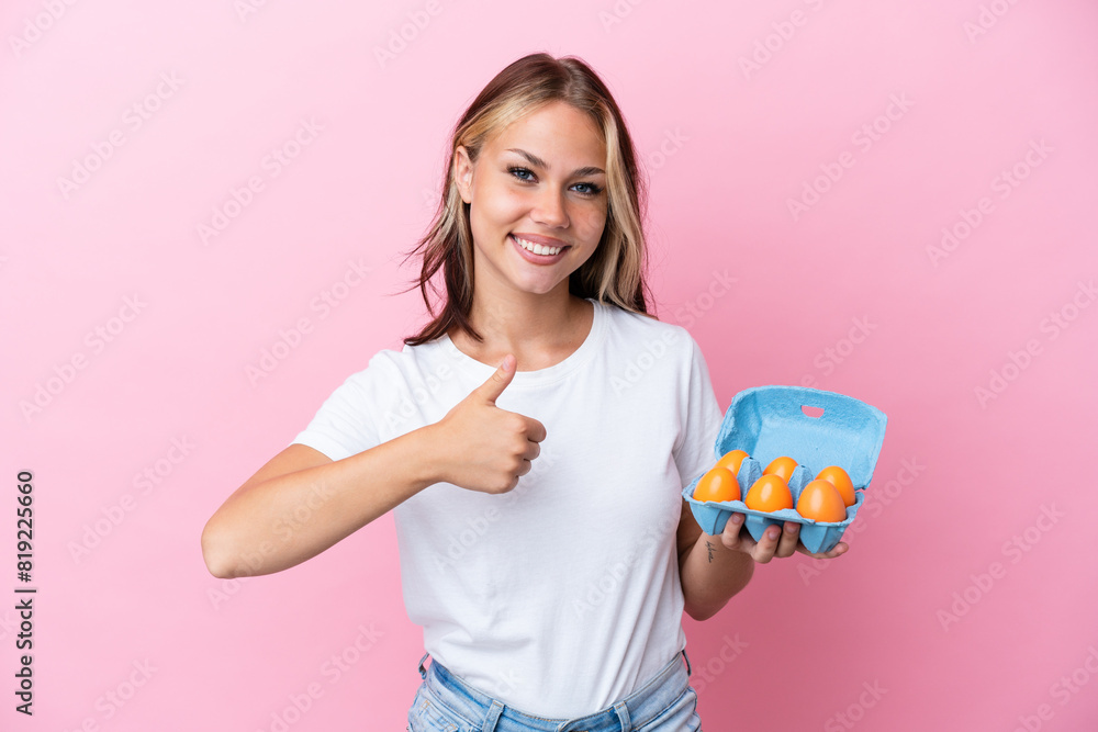 Young Russian woman holding eggs isolated on pink background giving a thumbs up gesture Young Russian woman holding eggs isolated on pink background giving a thumbs up gesture