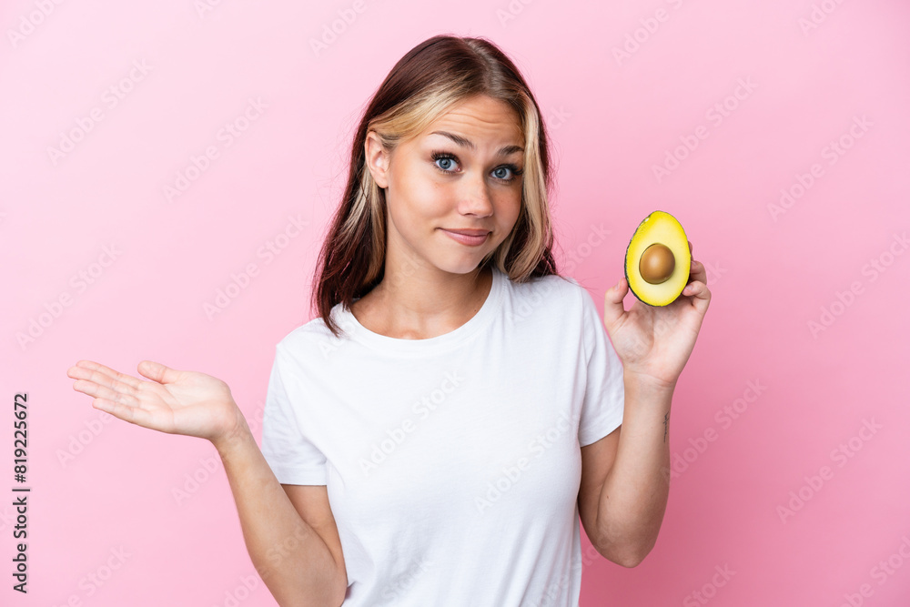 Young Russian woman holding avocado isolated on pink background having doubts while raising hands Young Russian woman holding avocado isolated on pink background having doubts while raising hands