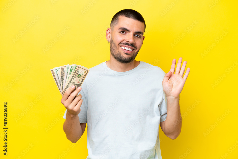 Young caucasian man taking a lot of money isolated on yellow background saluting with hand with happy expression Young caucasian man taking a lot of money isolated on yellow background saluting with hand with happy expression