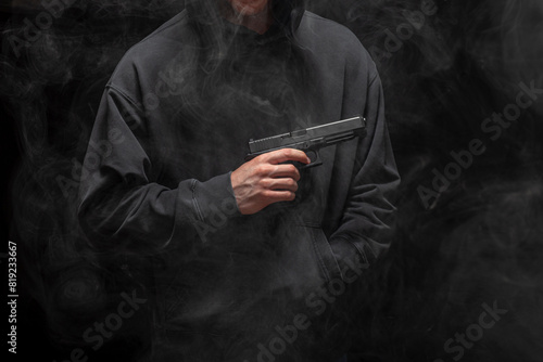 Young man in a black hoodie holding a pistol on a smokey black background