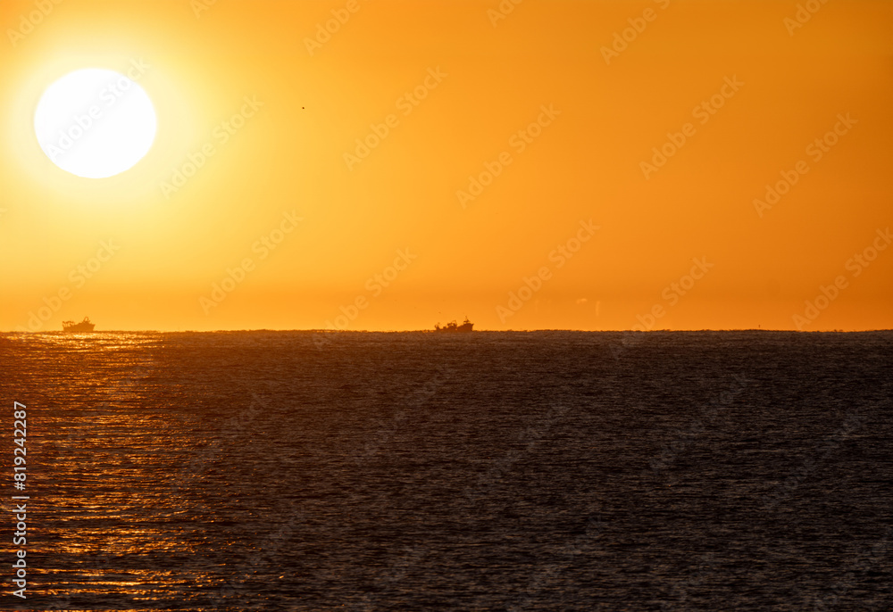 Yellow dawn sun emerging from the retro Mediterranean Sea horizon illuminating two fishing boats in silhouette heading out to catch shrimp, under an orange sky. Yellow dawn sun emerging from the retro Mediterranean Sea horizon illuminating two fishing boats in silhouette heading out to catch shrimp, under an orange sky.