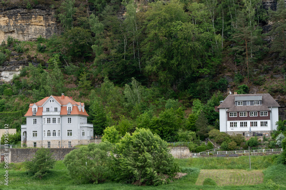 Obraz premium Two houses are on a hillside with a lush green forest in the background. The houses are white and brown, and they are situated on a hill with a view of the forest