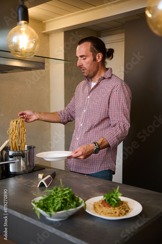 Male chef adding fresh green arugula leaves to Italian pasta with tomato sauce, garnishing the dish. A man preparing spaghetti for dinner in home kitchen. People and culinary concept. Italian cuisine