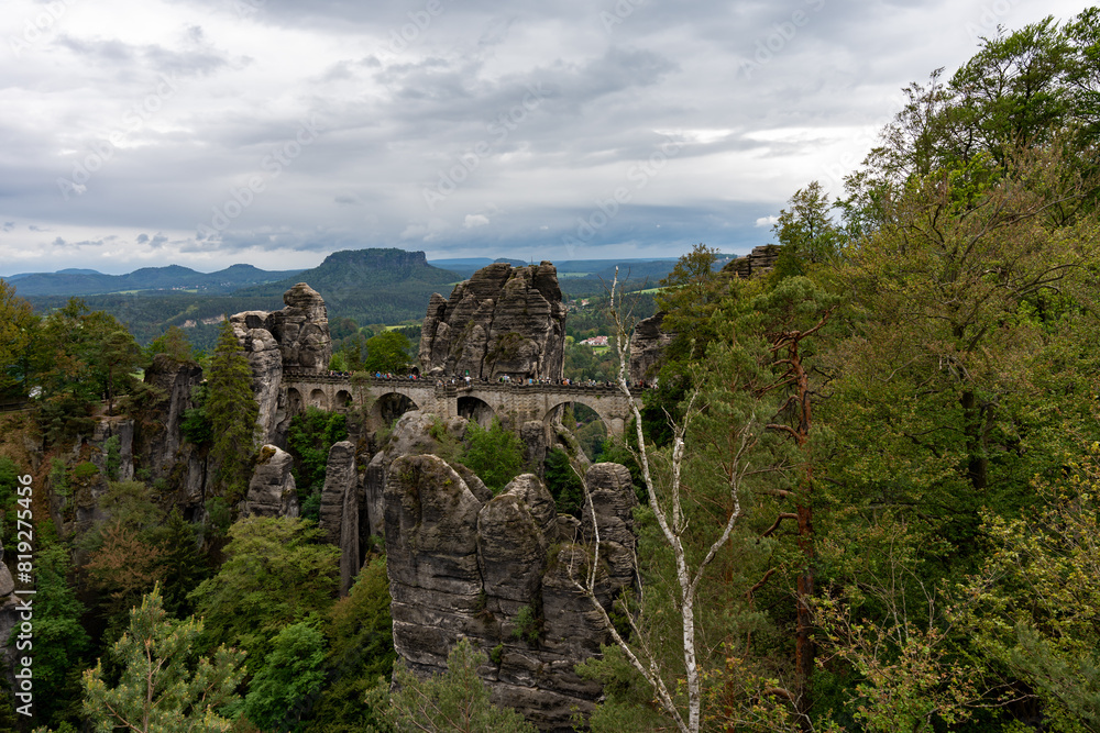 Naklejka premium A mountain range with a bridge in the middle. The bridge is surrounded by trees and the mountains are covered in clouds