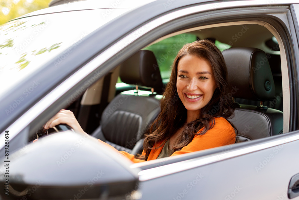 Happy European young lady driving car and enjoying road trip, looking at vehicle mirror with opened window and smiling
