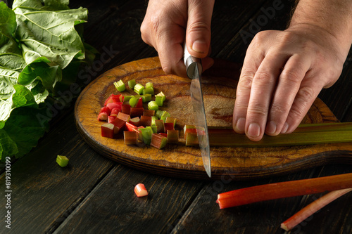 Male hands using a knife to cut rhubarb stalks on a wooden board for preparing a vegetarian dish at home. Peasant food