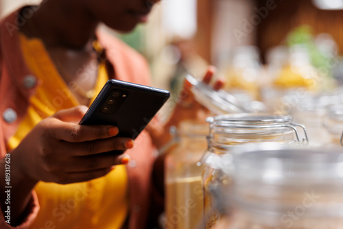 In this close-up image, black woman holds mobile device while checking out the items in the glass containers at the bio-food store. Selective focus on smartphone grasped by an african american female.