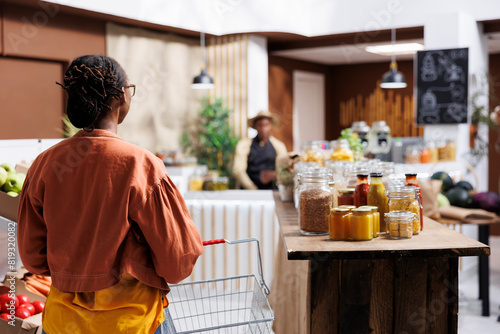 Black woman explores grocery store, browsing fresh, locally grown produce and sustainable packaging. Friendly seller provides excellent customer service in environmentally conscious supermarket.