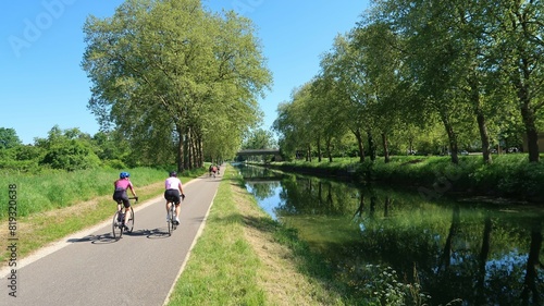 Cyclotourisme au bord du canal de Bourgogne à Dijon, en Côte d’Or, paysage de nature avec des arbres et deux cyclistes roulant sur une piste cyclable au bord de l'eau (France)
