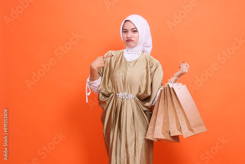 young asian girl sullenly carrying a brown paper bag containing shopping items while pointing at it wearing a kaftan Muslim dress with a hijab. Concept of fashion, transaction, promotion photo