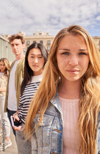 Vertical. Focus on serious young blonde woman looking at camera on street group multiracial friends in background. Confident people together posing portrait. Gen z colleagues sightseeing on vacation
