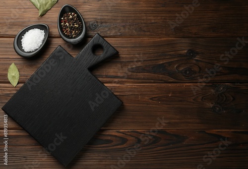 Black cutting board, salt, pepper and bay leaves on wooden table, flat lay. Space for text photo