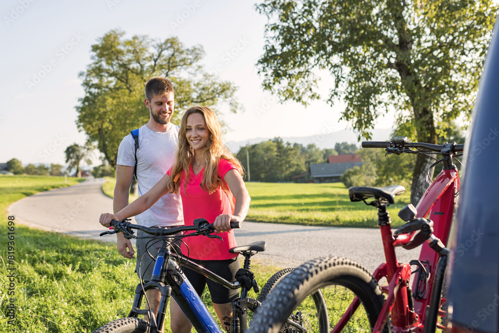Young man is unloading electric mountain bikes, for himself and his girlfriend, lifting them from the hitch rack on the vehicle. Young man is unloading electric mountain bikes, for himself and his girlfriend, lifting them from the hitch rack on the vehicle.