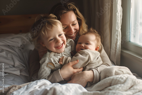 Laughing mom cuddling happy young son and baby on bed near window