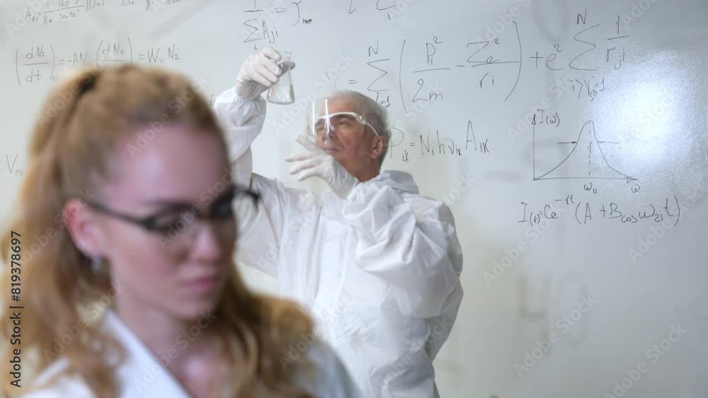 A woman chemist writes a formula on glass. An elderly Caucasian man in a protective suit is doing tests. A woman chemist writes a formula on glass. An elderly Caucasian man in a protective suit is doing tests.
