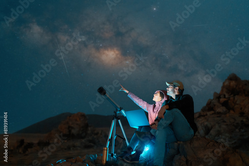 heterosexual couple sitting in the desert watching the stars and Milky Way next to a telescope, stargazing and exploration concept	