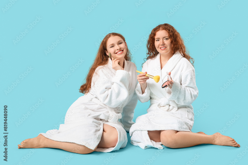 Happy redhead sisters in bathrobes with massage tool and makeup supplies on blue background