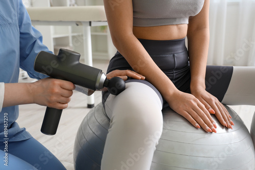 Female physiotherapist massaging young woman's thigh with percussive massager on fitball in rehabilitation center, closeup photo