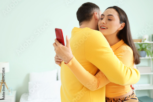 Young woman with mobile phone hugging her husband at home. Cheating concept
