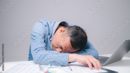 The tired businessman, a sleepy man with a headache, slumps over his desk in the office, struggling to stay awake.
