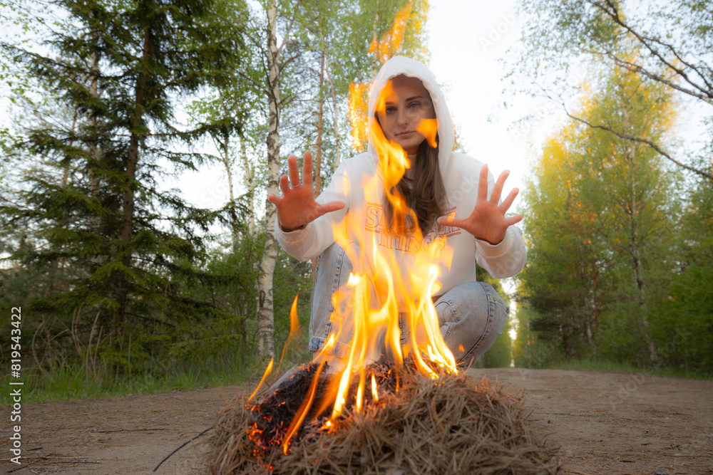 woman in a white hoodie crouches on a dirt forest path, intensely focused on a small fire she has started. Tall birch trees with lush green foliage line the path, adding to the serene yet intense woman in a white hoodie crouches on a dirt forest path, intensely focused on a small fire she has started. Tall birch trees with lush green foliage line the path, adding to the serene yet intense