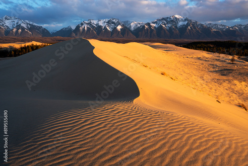Chara Sands, Kodar nature reserve.Ripples in the sand dunes are highlighted by the warm glow of a rising sun. IIn the background, majestic snow-capped mountains stand tall against a partly cloudy sky, photo