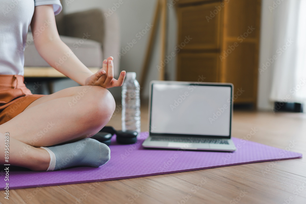 Fototapeta premium Young Asian woman at home doing yoga in front of open laptop, repeating instructions by professional online fitness trainer.