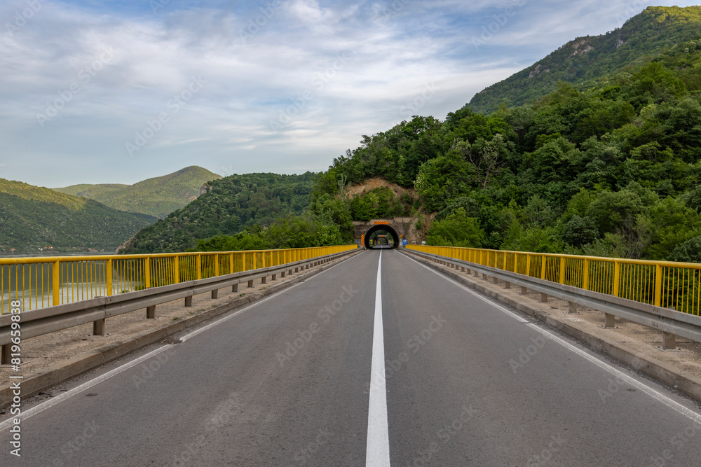 A straight asphalt road with yellow guardrails on both sides leads towards a tunnel entrance, surrounded by mountains and lush green trees under a clear sky... A straight asphalt road with yellow guardrails on both sides leads towards a tunnel entrance, surrounded by mountains and lush green trees under a clear sky...