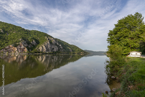 Wallpaper Mural  captures a serene river scene with a cabin and boat on the grassy bank, set against a backdrop of cliffs and mountains. The calm water reflects the blue sky and greenery, creating a peaceful  Torontodigital.ca