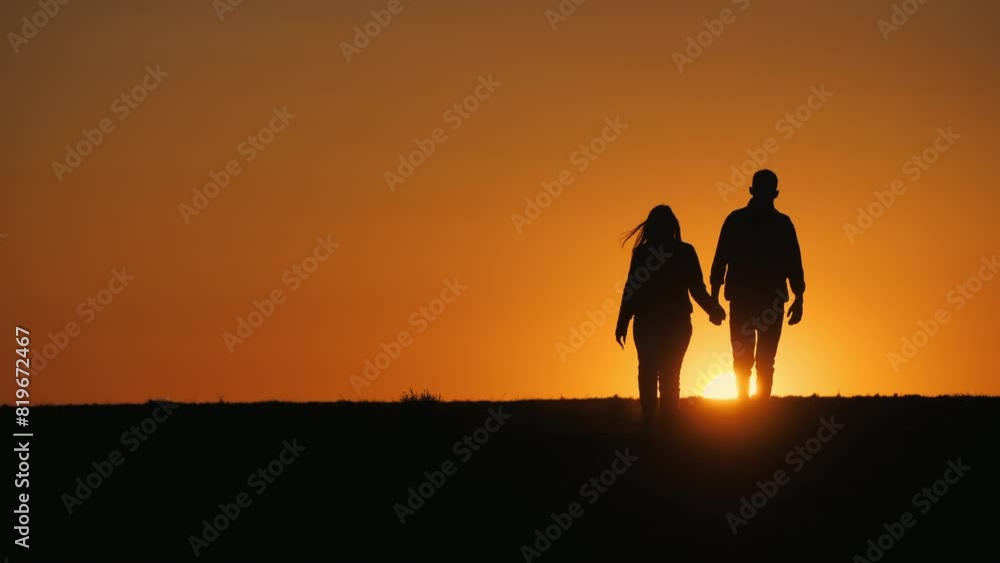 Against the backdrop of the setting sun, a young couple walks hand in hand across a field. Against the backdrop of the setting sun, a young couple walks hand in hand across a field.