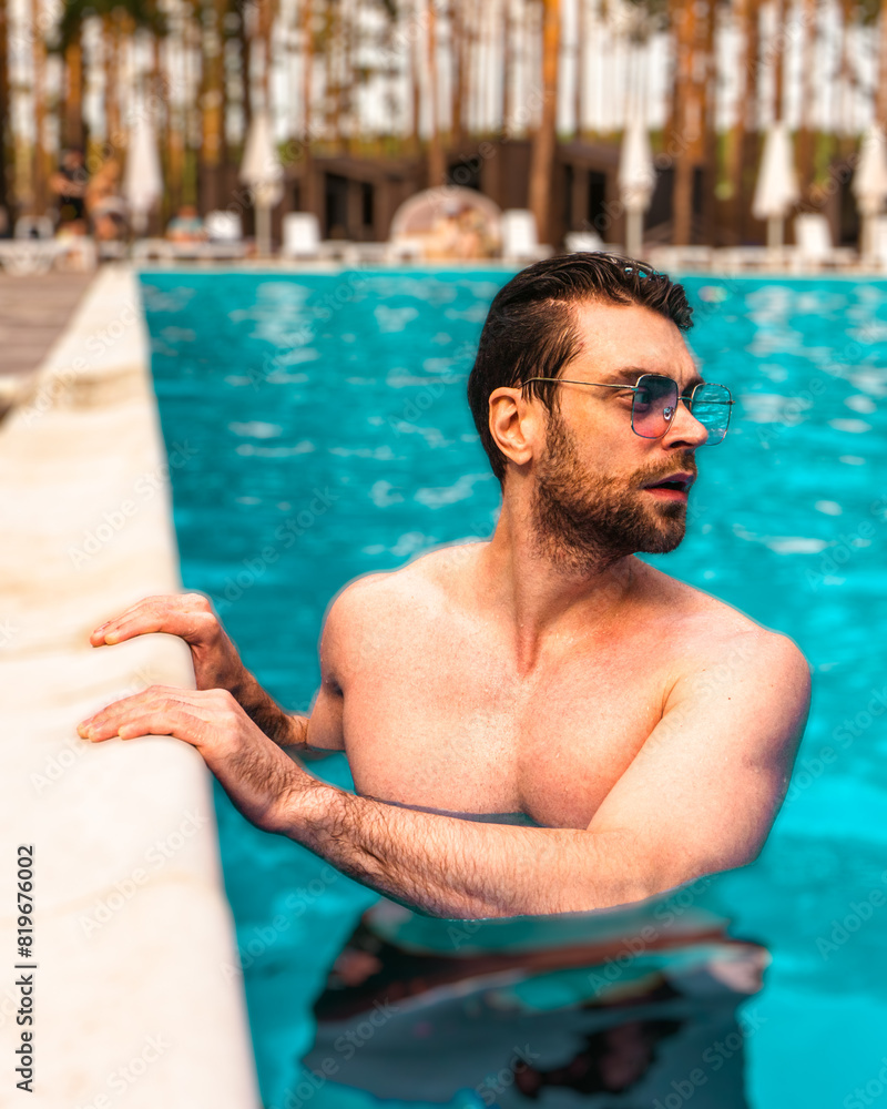 Young man standing in the water while having rest in the swimming pool outdoors, looking away. Summertime, holidays, lifestyle concept