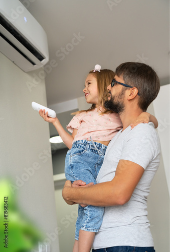 Happy little girl holds remote control and turn on air conditioner sitting in fathers hands at home