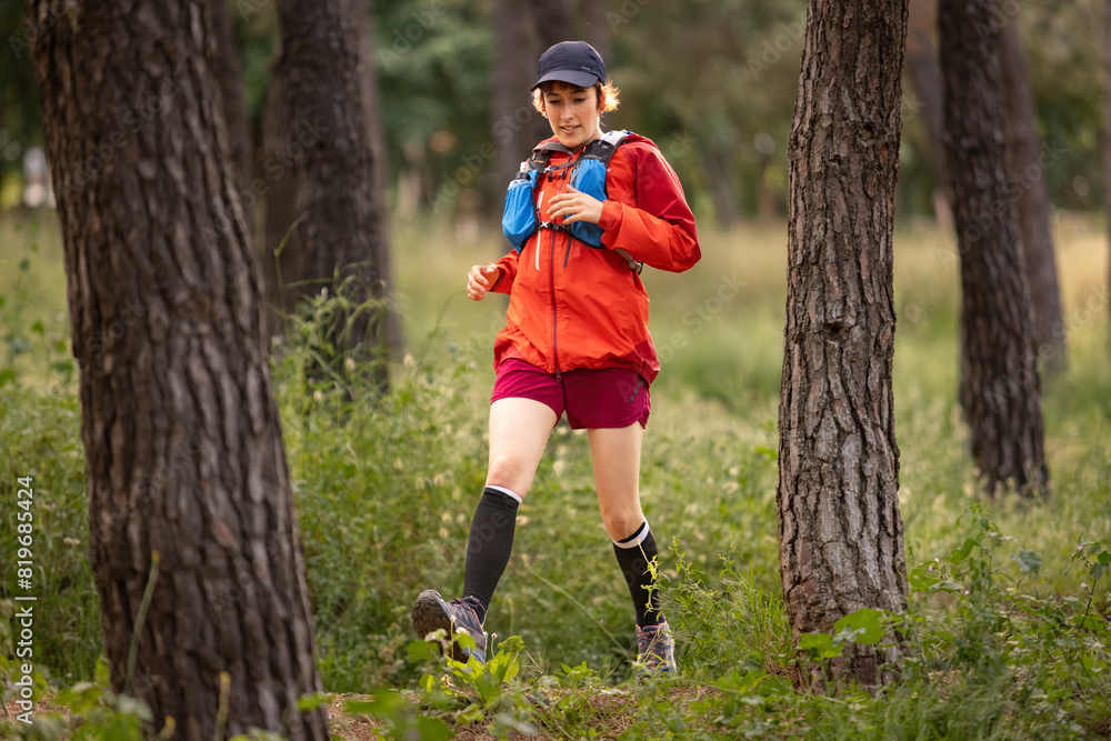 A woman is running through a forest wearing a red jacket and black hat. She is wearing black socks and a backpack A woman is running through a forest wearing a red jacket and black hat. She is wearing black socks and a backpack