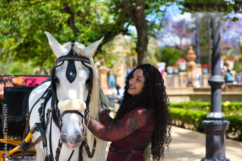 Obraz premium latin woman, brunette with curly hair, young and beautiful next to an elegant white horse pulling a horse and carriage. The woman is in seville on holiday and enjoys the sunshine of southern spain.