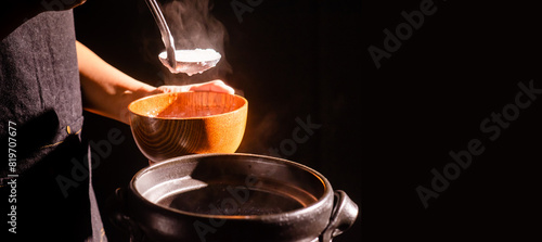 The cook was cooking rice porridge in a boiling clay pot with steam coming out and using a ladle to scoop the rice porridge into a wooden bowl. photo